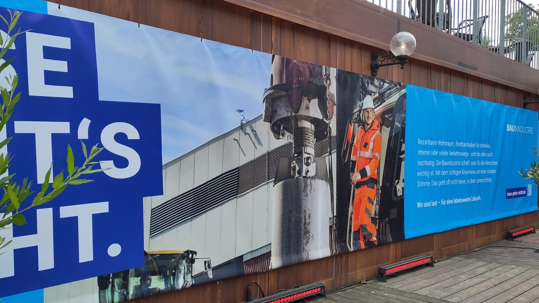 Ansicht eines großflächigen Banners, das einen Bauarbeiter auf einer Maschine und den Kampagnenclaim zeigt, an einem Gebäude.